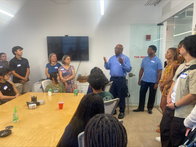 An alum who is standing speaks to a group of alums and students who are standing or sitting at a table