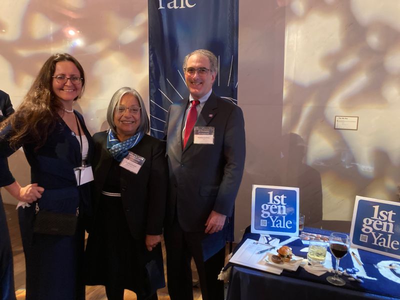 Three alums stand to the side of a table with 1stGenYale materials