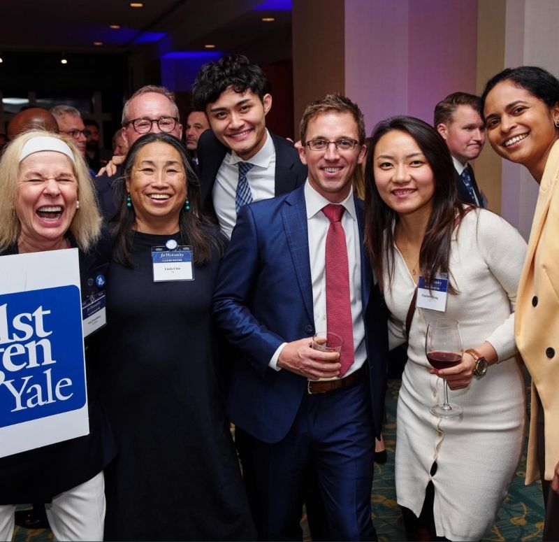 Seven alums standing, laughing and smiling, and holding a 1stGenYale sign
