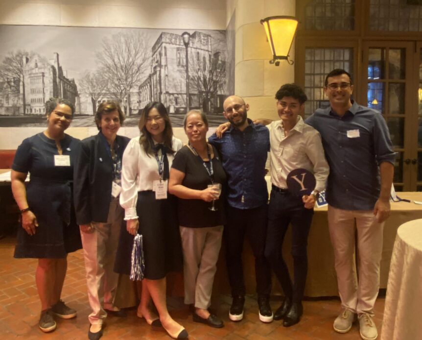 Seven Yale alums stand in a row in front of a table and a drawing on the wall of the Yale campus