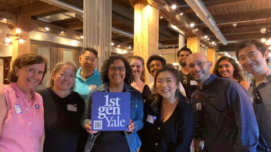 Eleven people stand inside a large room with columns and smile at the camera and hold a 1stGenYale sign