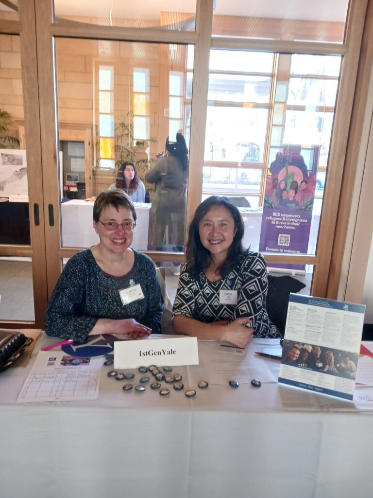 Two women are seated at a table with 1stGenYale flyers, buttons, and signs in front of them and in back behind a glass divider a student talks with a person representing a nonprofit employer