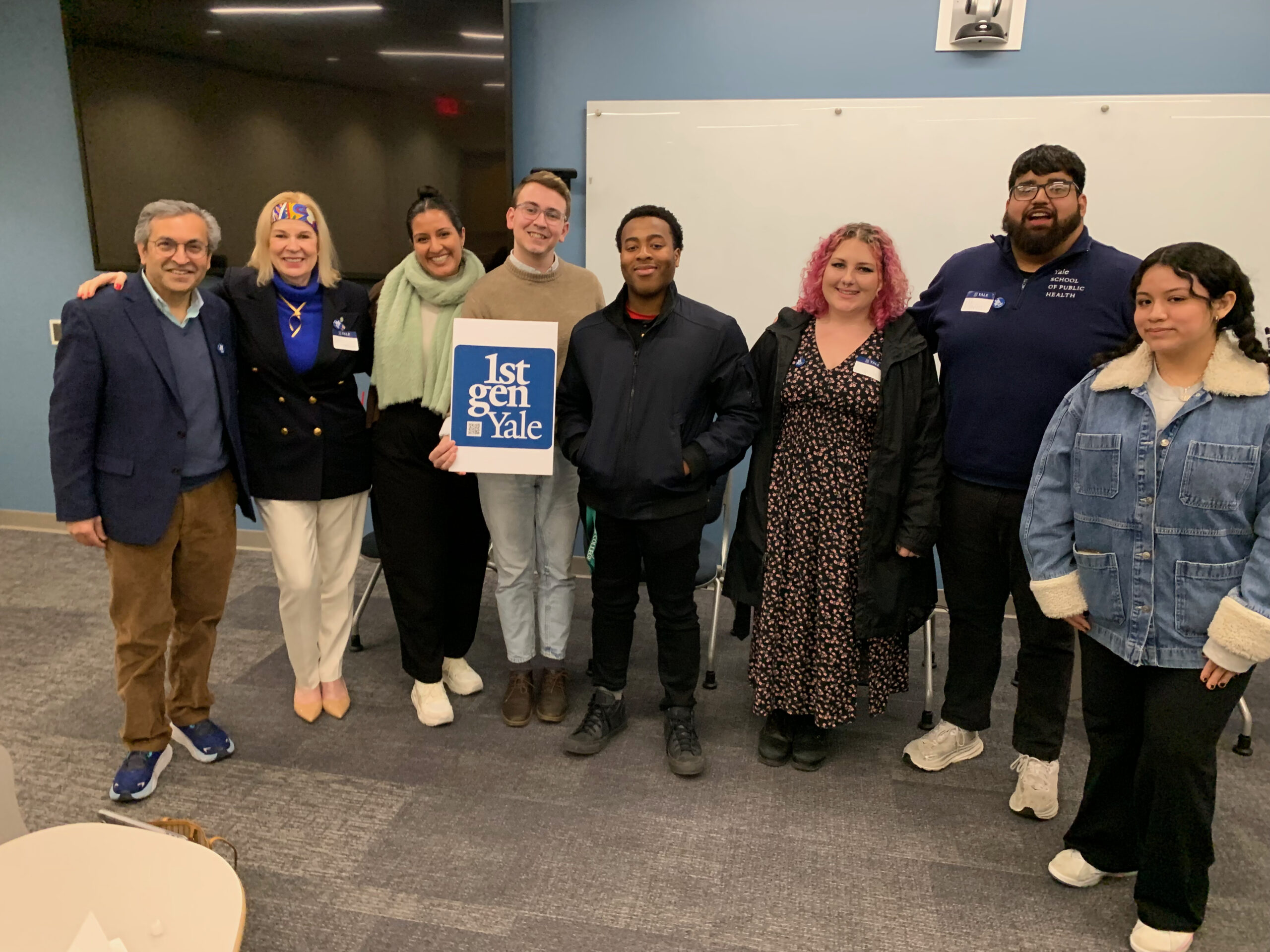 Eight people stand facing the camera and hold a 1stGenYale sign