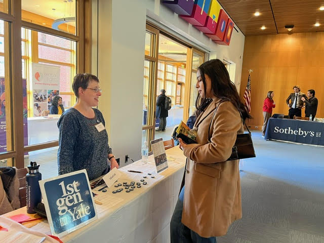 A student stands and talks with a woman at a table with 1stGenYale signs and flyers