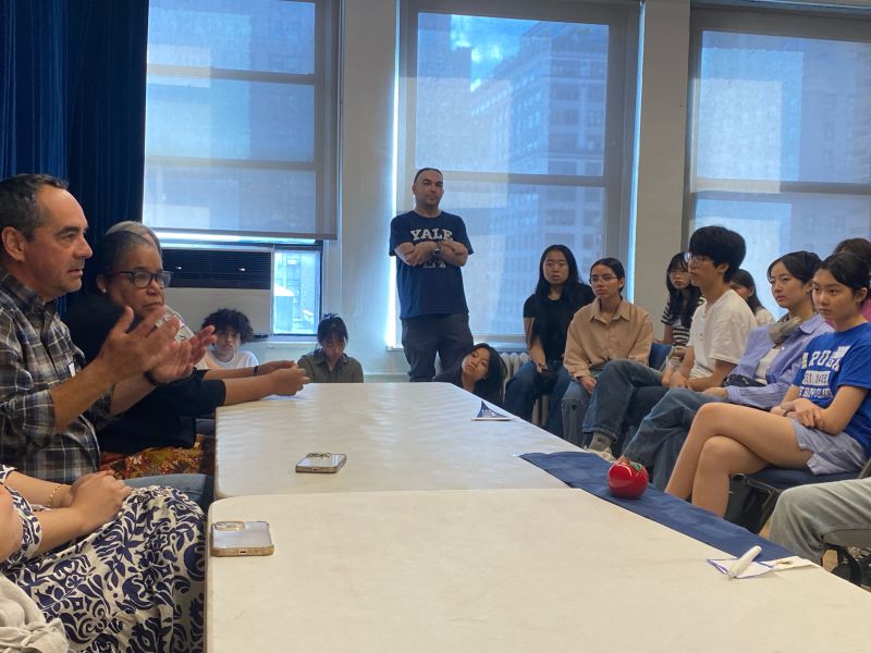 An alum stands in front of a window while alums who are seated talked with students
